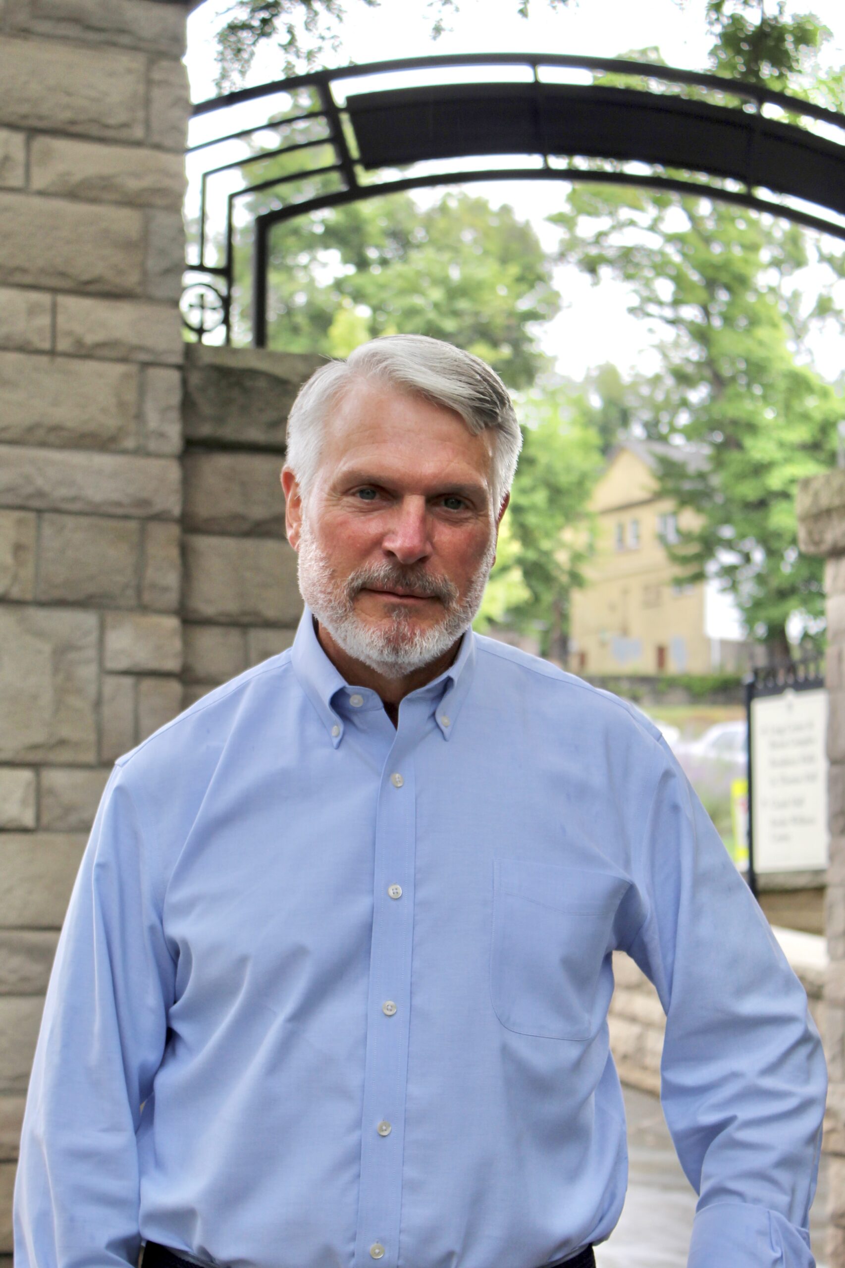 A man with gray hair and a short beard stands outdoors wearing a light blue button-down shirt. He is in front of a stone archway with trees and buildings visible in the background.