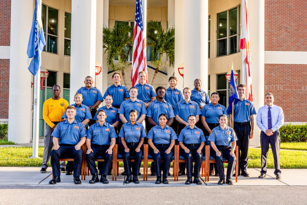 Group portrait of Indian River State College law enforcement academy cadets in blue uniforms, arranged in three rows outside a columned campus building with palm trees. U.S. and Florida flags stand behind; an instructor in yellow is at left and a staff member in a tie is at right.