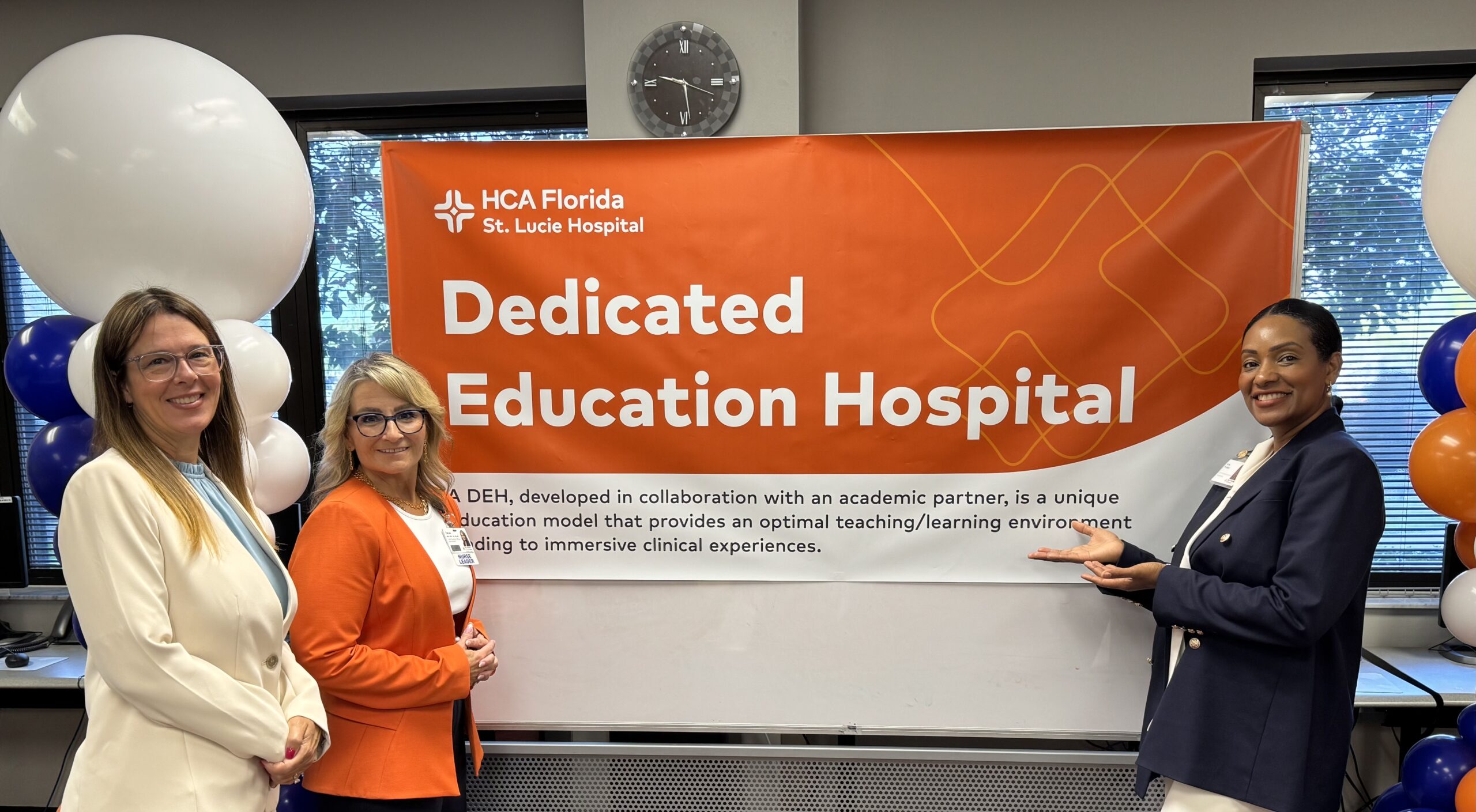 Three women stand in front of an orange banner reading “HCA Florida St. Lucie Hospital Dedicated Education Hospital.” The banner describes a collaboration with an academic partner to create an immersive clinical education model. The women are smiling, dressed in professional attire, and surrounded by blue, white, and orange balloons.