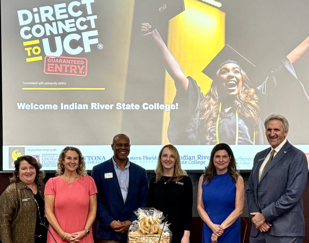 Six representatives from Indian River State College and the University of Central Florida stand side by side in front of a large screen displaying the “DirectConnect to UCF – Guaranteed Entry” logo. From left to right: Adrienne Frame, Elizabeth Gaskin, Calvin Williams, Kimberly Hardy, Heather J. Belmont, and John Buckwalter. All are smiling and dressed in business attire, gathered to celebrate IRSC’s launch into DirectConnect to UCF.