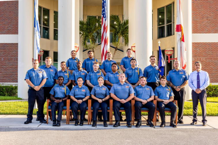 Group portrait of Indian River State College law enforcement academy cadets in blue uniforms, posed in three rows outside a columned campus building. The U.S. flag is centered, with campus and Florida/academy flags flanking the group; a staff member in a dress shirt and tie stands at far right.