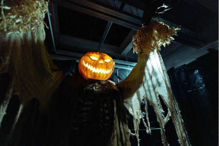 A spooky Halloween decoration featuring a scarecrow figure with a carved jack-o'-lantern pumpkin head. The pumpkin has a menacing grin with jagged triangular teeth and glowing yellow eyes. The scarecrow is dressed in tattered, weathered clothing and positioned with one arm raised upward. The scene is set in what appears to be an indoor space with exposed wooden ceiling beams and structural elements visible in the dim lighting, creating an eerie haunted house atmosphere.