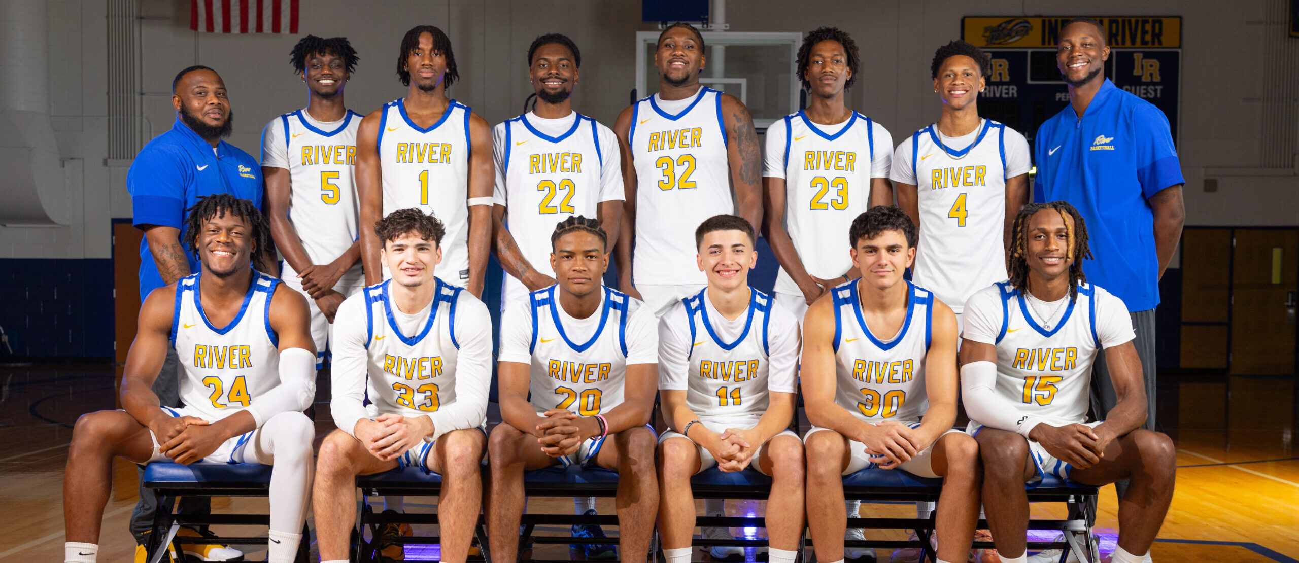 A group of 13 basketball players and 2 coaches from Indian River State College pose for a team photo in the gym, wearing white and blue “RIVER” uniforms and smiling confidently.