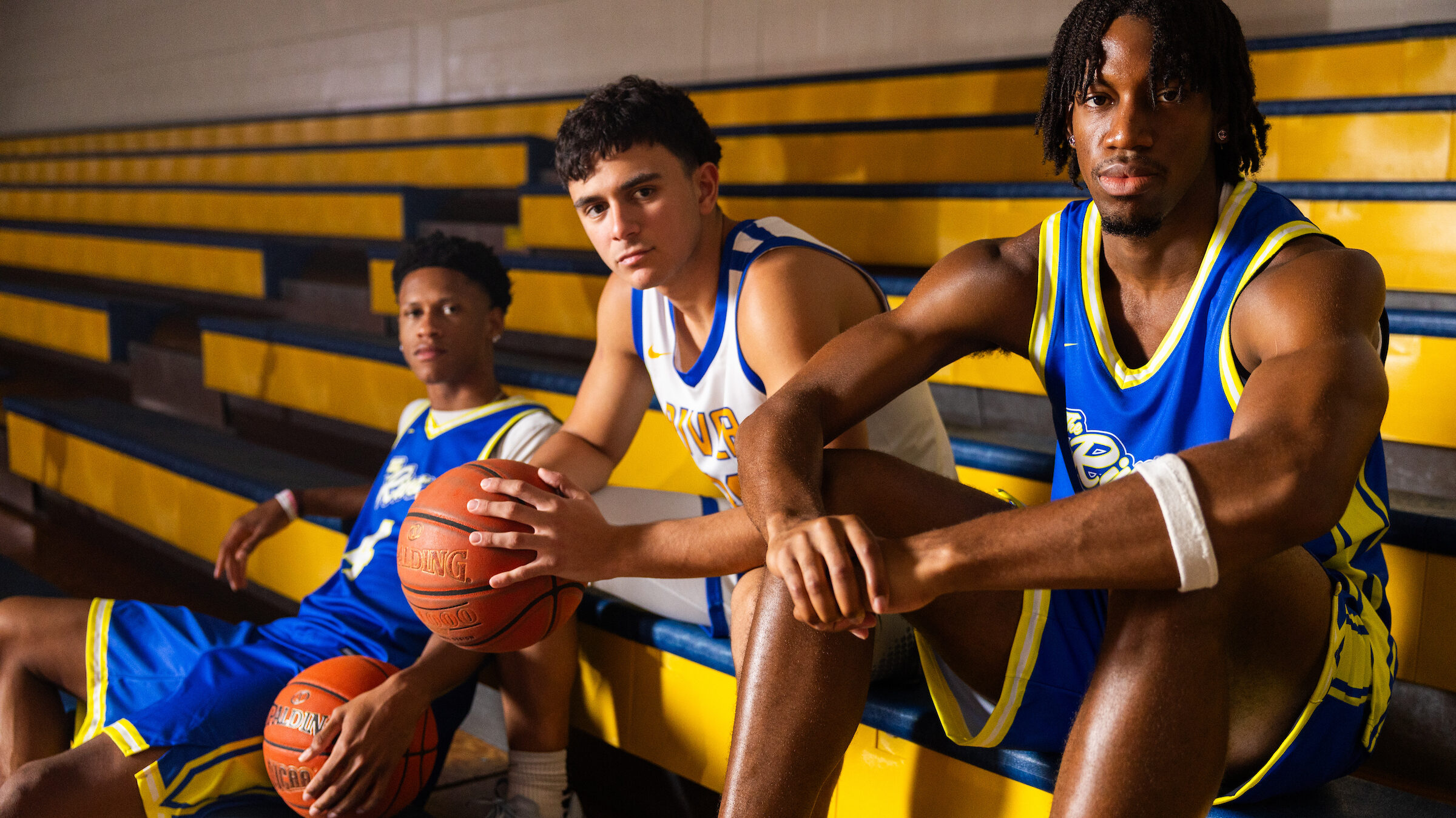 Three Indian River State College basketball players in blue and white uniforms sit on yellow bleachers holding basketballs, showing focus and confidence before the new season.