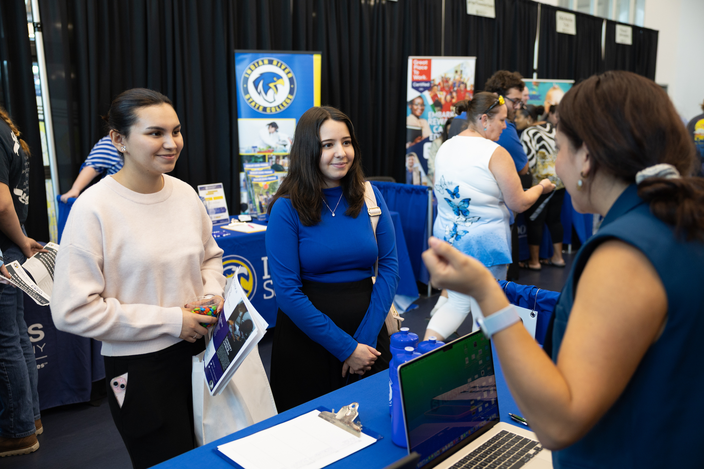 Two students speak with a representative at an Indian River State College career expo. The representative gestures while standing behind a table with college materials and a laptop. Banners with the IRSC logo and program information are visible in the background.
