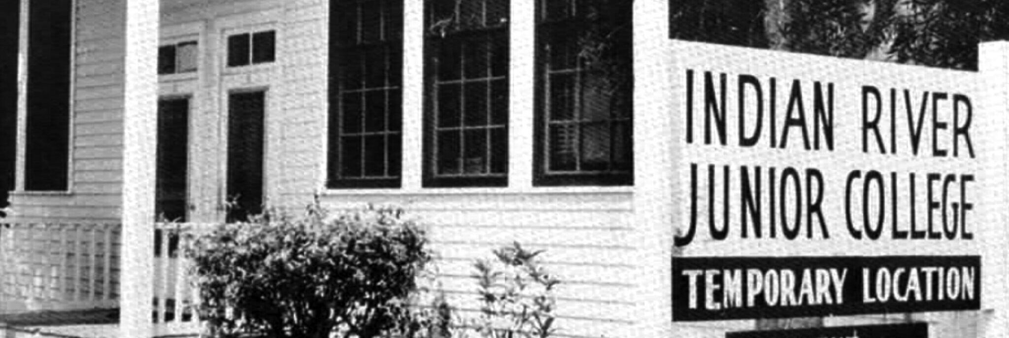 A black and white photo of a while building and a sign reading "Indian River Junior College - Temporary Building" with part of a tree and bushes.