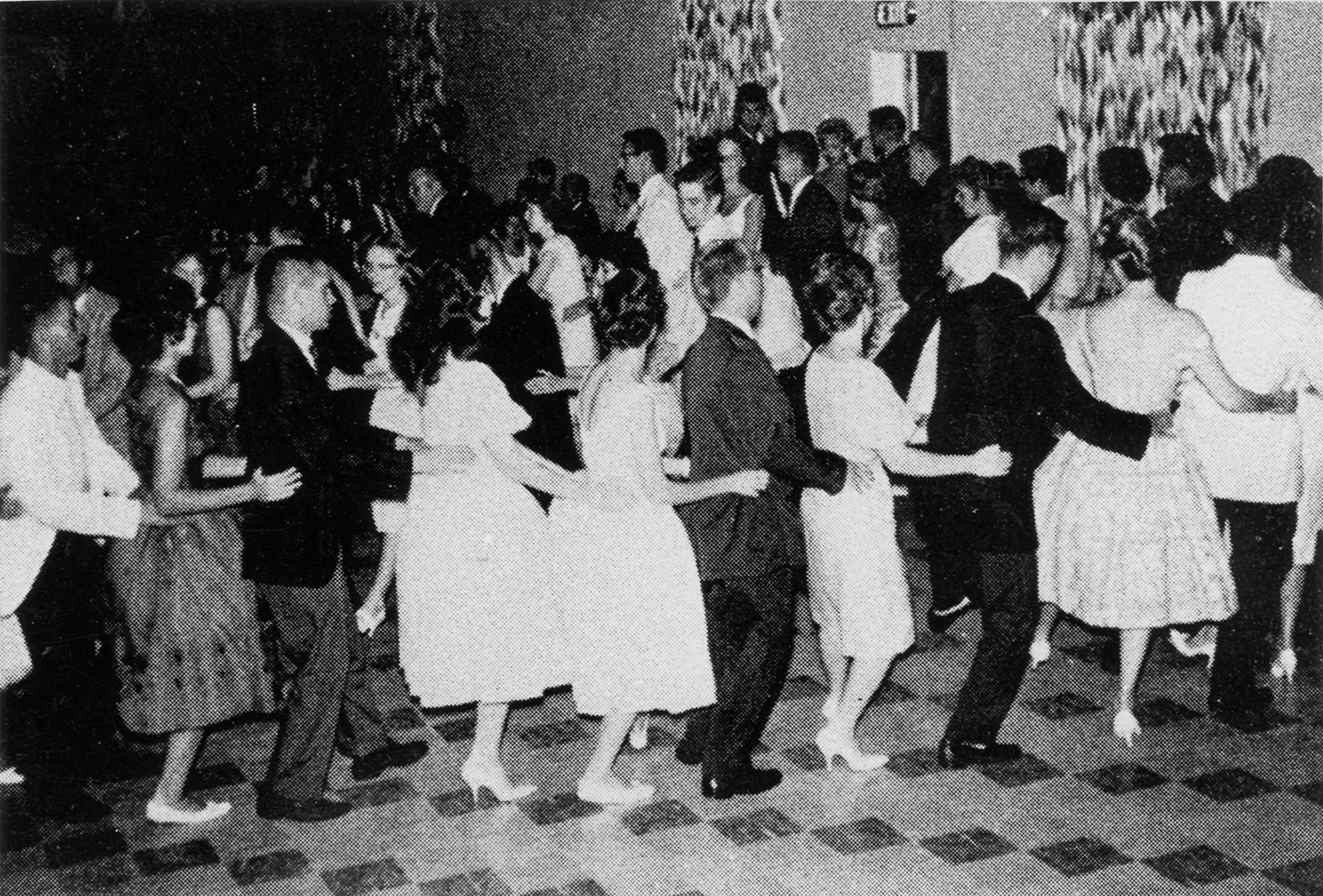 A black and white photo of men in suits and women in dresses doing the conga dance.