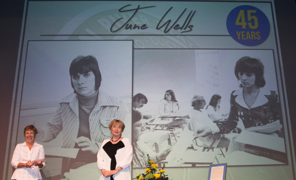 A photo of two women on a stage of whom one is Professor June Wells, receiving an award for her years of service, in 2018.
