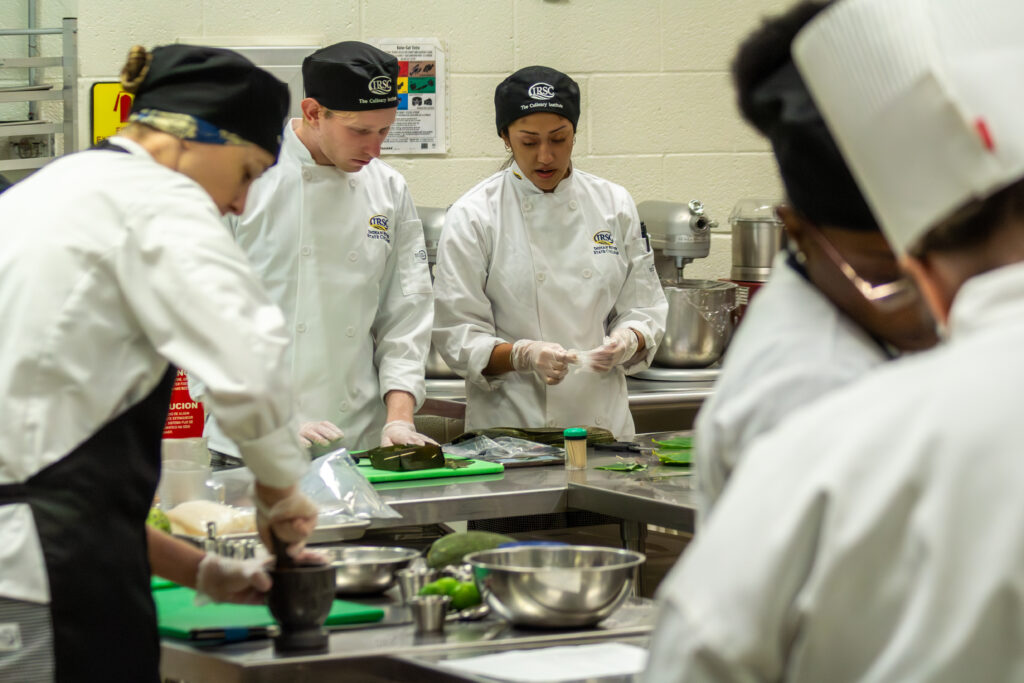 Five students gather around a prep table with food on it, as they prepare a menu in a kitchen.