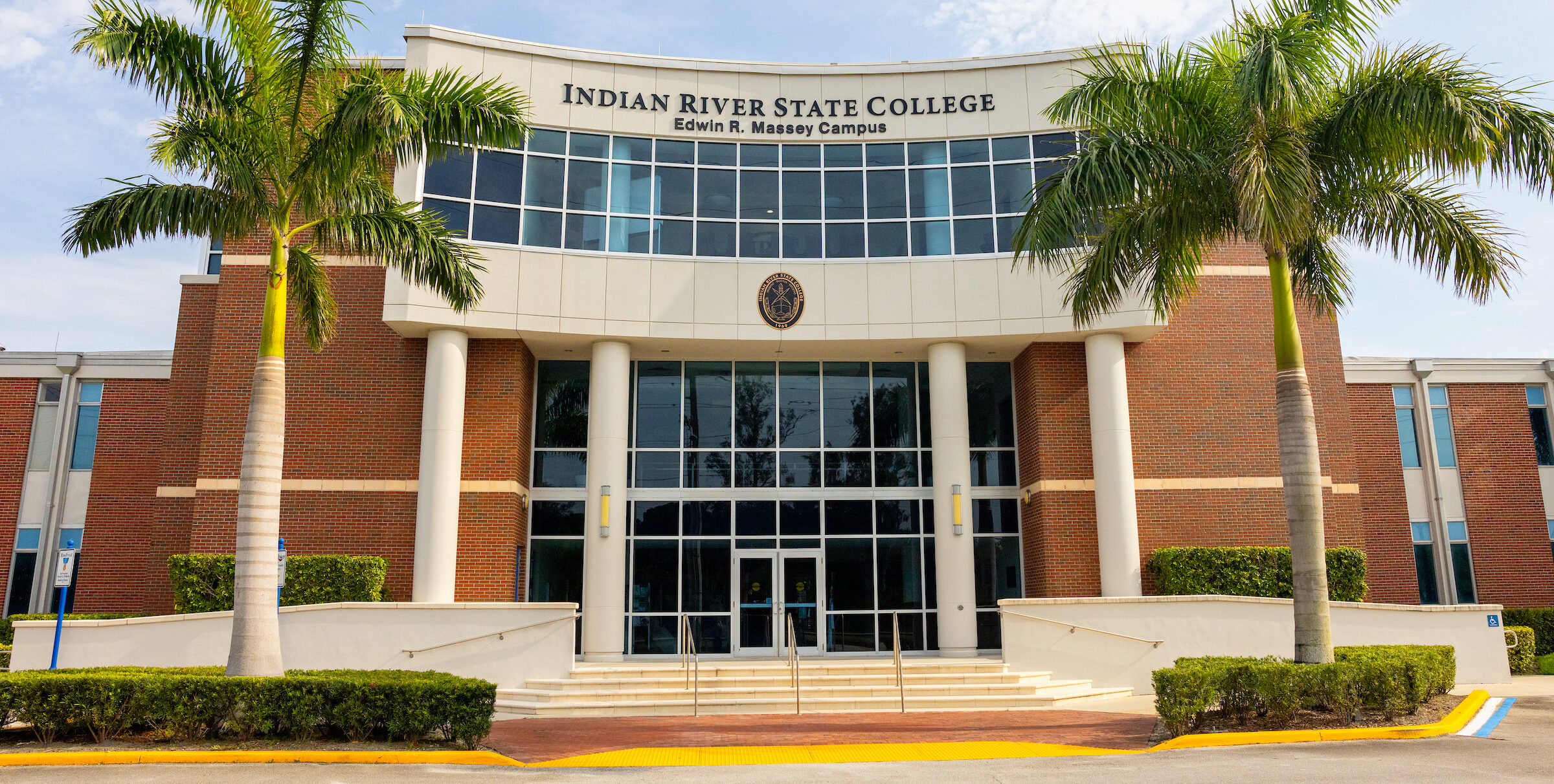 Front view of the Indian River State College Edwin R. Massey Campus building in Fort Pierce, Florida, featuring brick architecture, tall columns, and palm trees.
