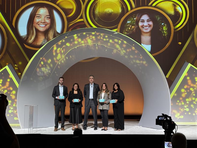 A photo of five people standing on a stage at a conference. The photo depicts an award presenter and four award recipients, including Jenna Bluedorn.