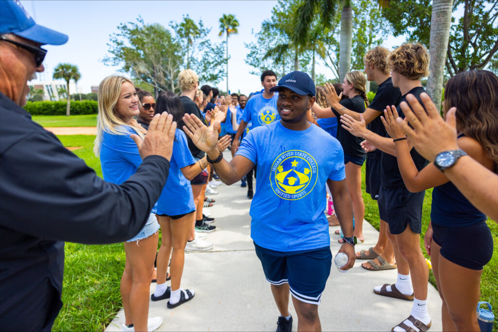 A group of Indian River State College students wearing blue “Unified Sports” shirts high-five each other along a sunny campus walkway lined with palm trees, smiling and cheering in a show of unity and school pride.