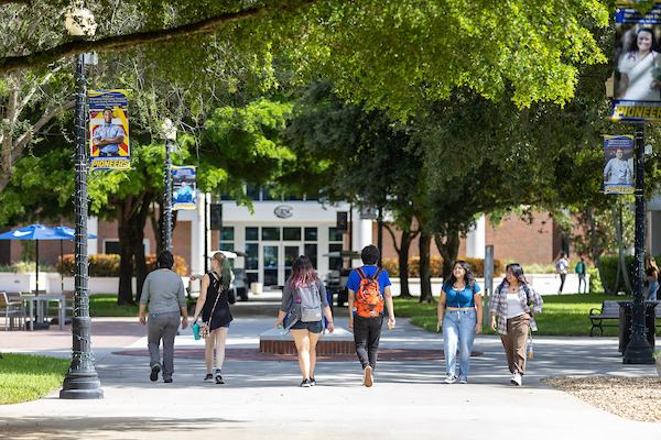 On a sunny day, several students walk along a tree-lined path, with an administration building in the background.