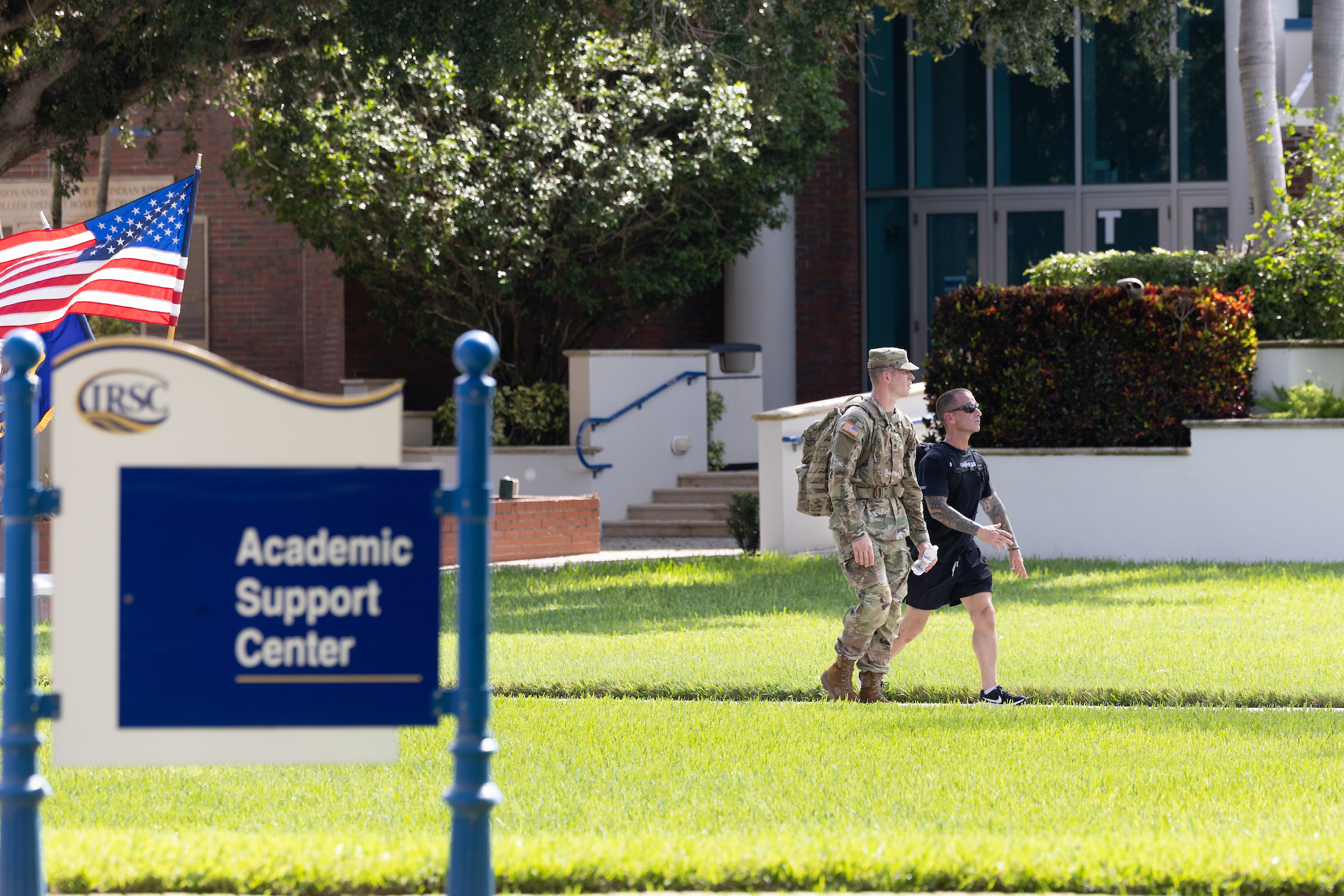Two individuals walk on the grounds of Indian River State College's main campus in Fort Pierce, Florida. An American flag waves atop a sign that reads "Academic Support Center."