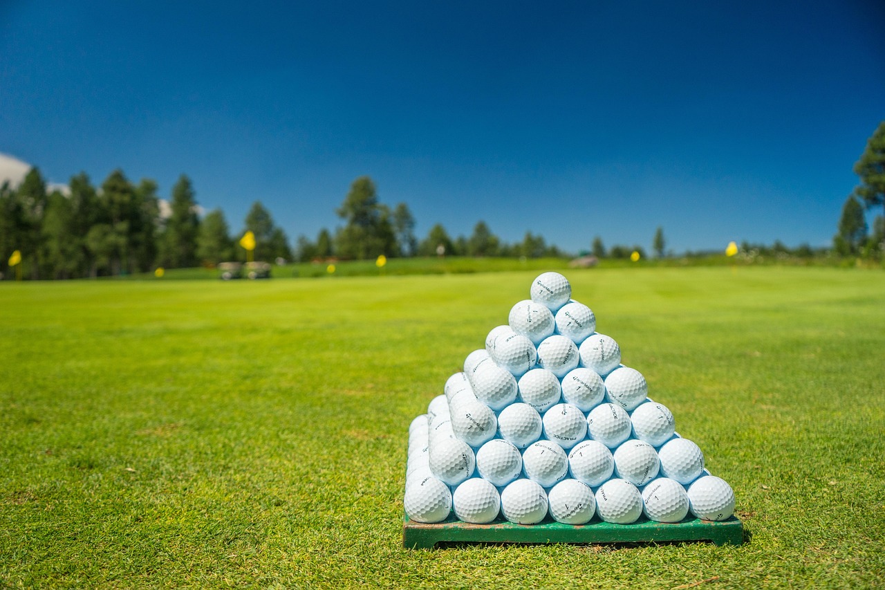 A pyramid of golf balls sits atop a golf course green, with trees in the background and blue sky.