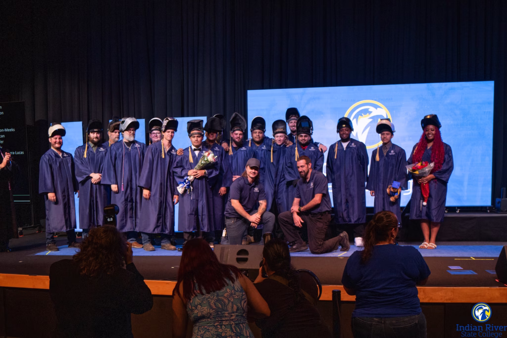 Large group of graduates in blue caps and gowns standing on stage with Indian River State College logo displayed on screen behind them. Two faculty or staff members in dark polo shirts kneel in front of the group. Several graduates hold bouquets of flowers. Audience members visible in foreground watching the ceremony.