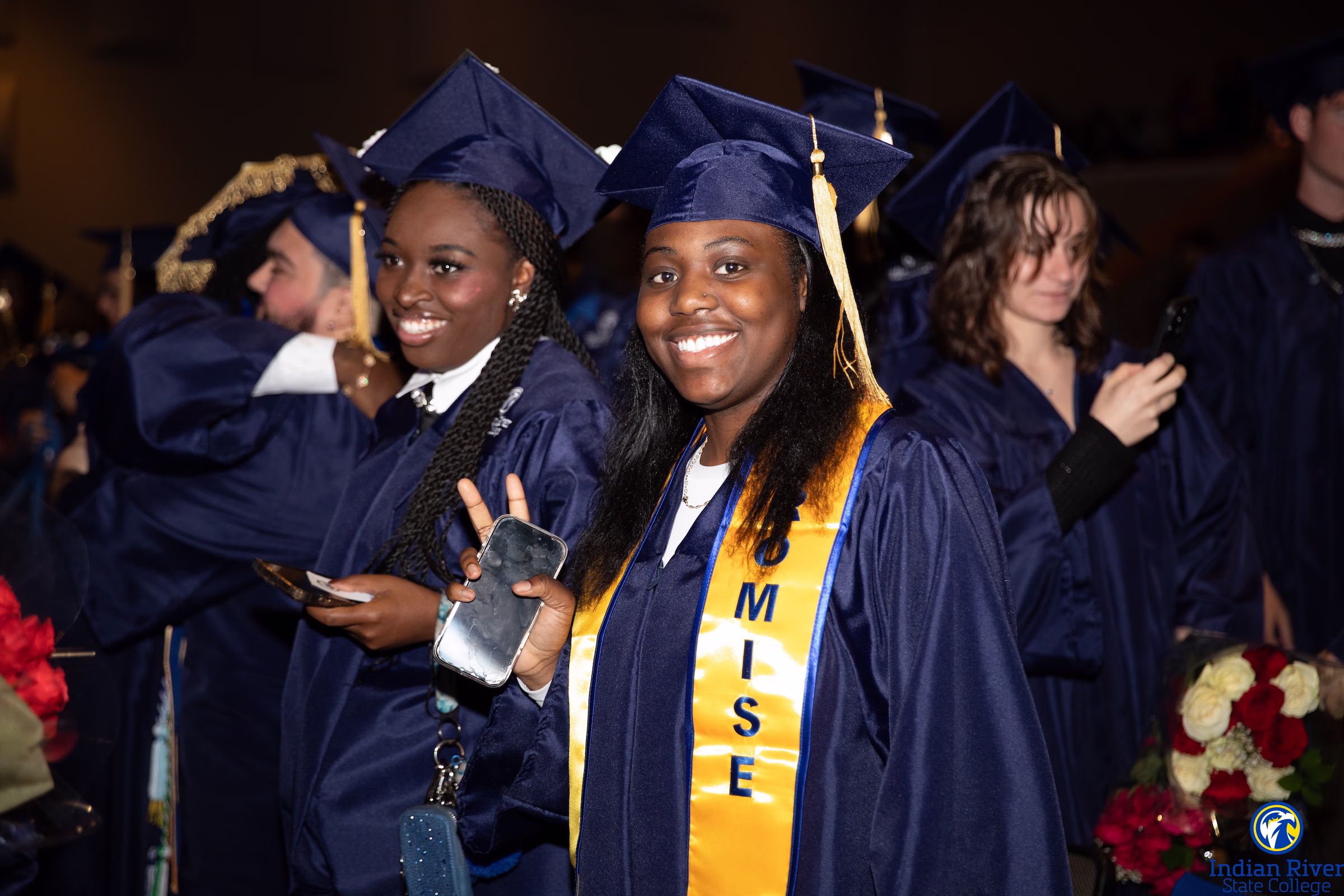 A woman wearing a blue graduation cap and gown, with a stole with the word "Promise" on it, smiles and waves. She is surrounded by fellow graduates.