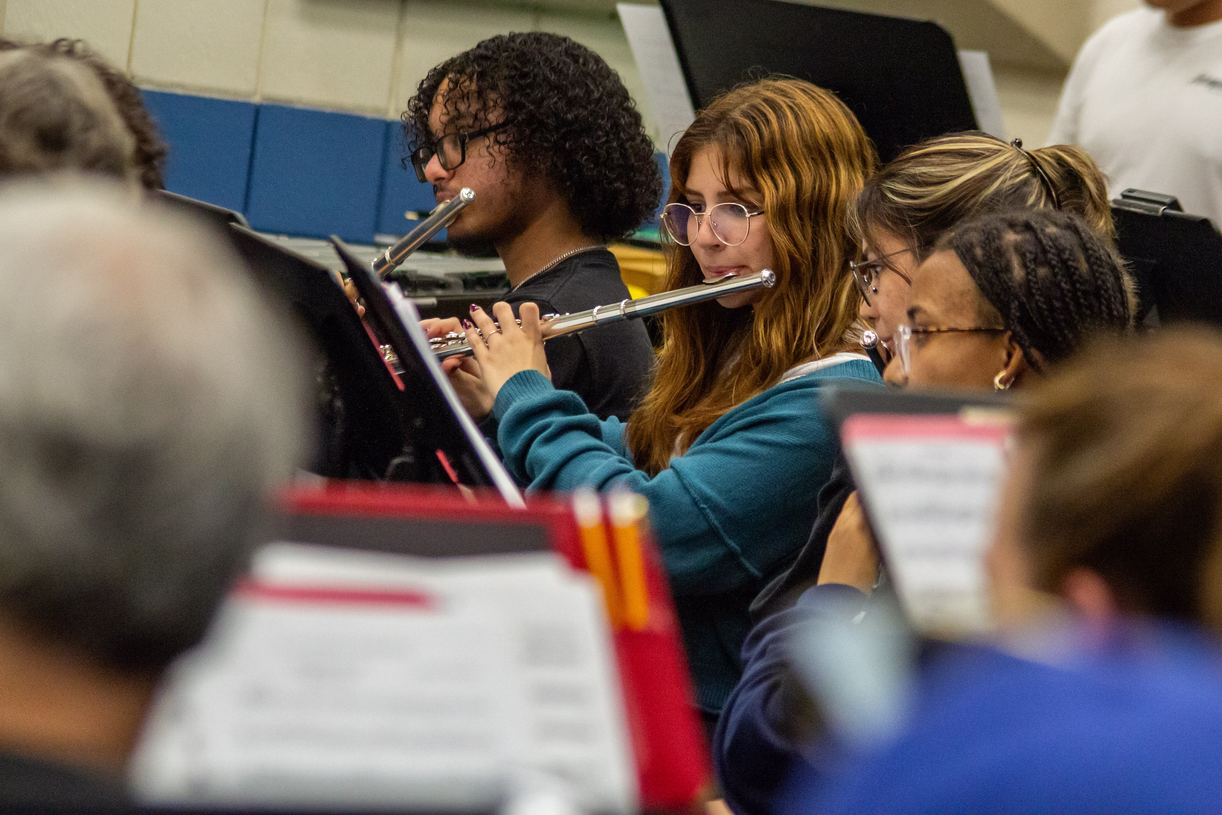Three flute players rehearsing in a band or orchestra setting. The musicians are seated with sheet music on stands in front of them. In the foreground center, a young woman with long brown hair and glasses wearing a teal sweatshirt plays her flute, flanked by two other flutists. Additional band members with music stands are visible in the blurred foreground and background.