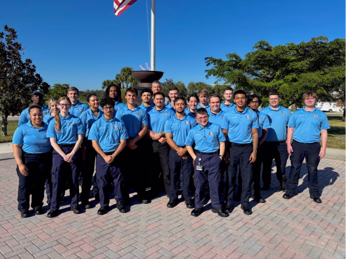 A group of approximately 30 young people in matching light blue polo shirts and dark navy pants standing together outdoors in front of an American flag and flagpole with an eternal flame monument, surrounded by palm trees and other vegetation on a sunny day.
