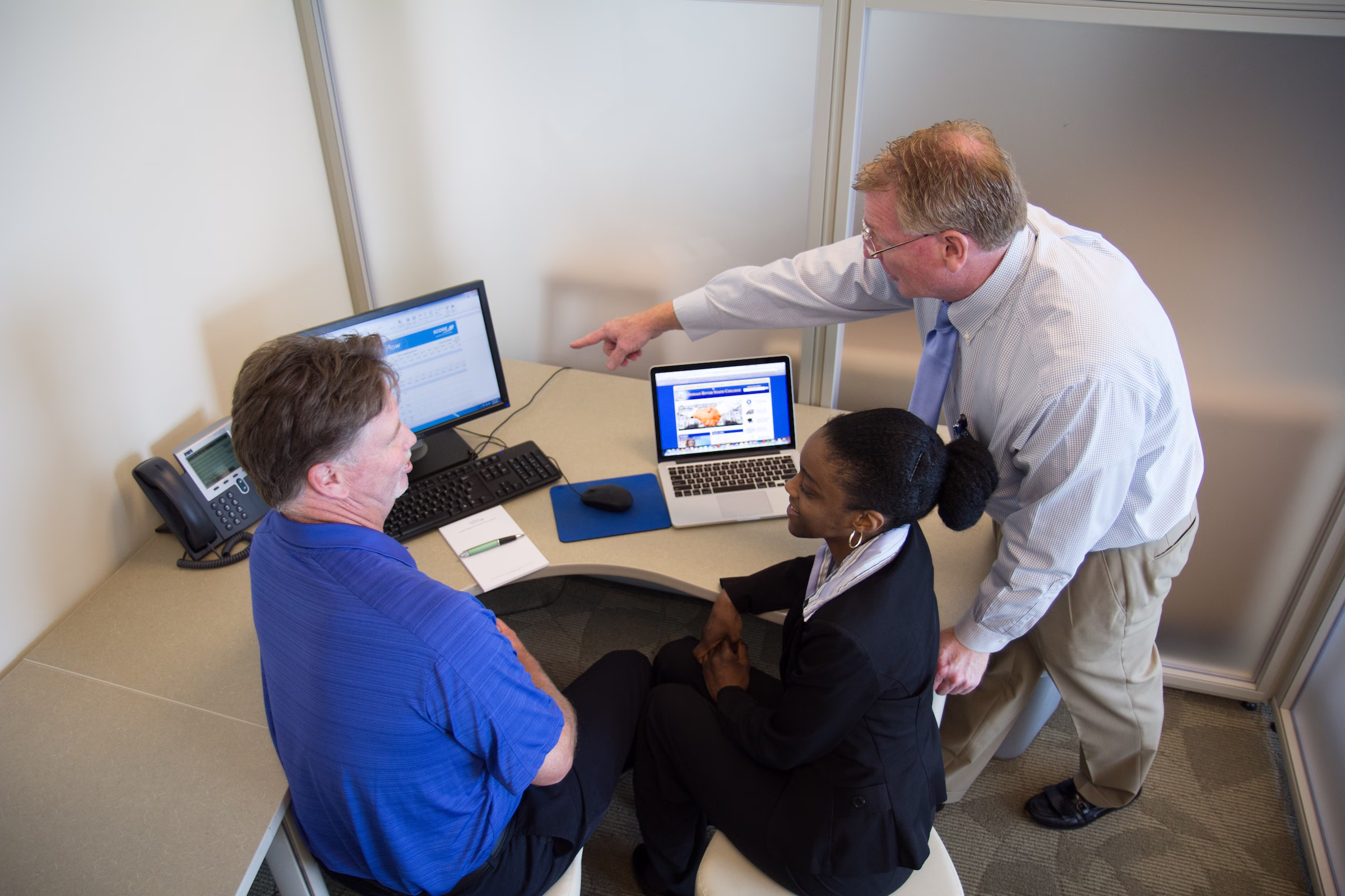 Three people collaborating in a small office workspace. A man in a white dress shirt and blue tie stands and points to a laptop screen while consulting with two seated individuals - a woman in a blue shirt and a woman in business attire. The desk has a desktop computer monitor, laptop, phone, notepad, and mouse pad. The setting appears to be a business consulting or training session.