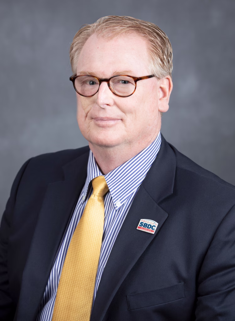 Professional headshot of a man with blonde hair wearing round tortoiseshell glasses, a dark suit jacket, blue and white striped dress shirt, and a gold tie. He has an SBDC (Small Business Development Center) pin on his lapel and is smiling at the camera against a gray background.