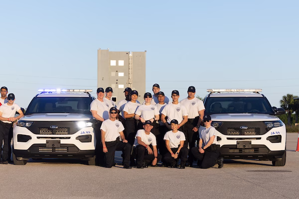 A group of approximately 15-20 public safety students in white uniform polo shirts pose together in front of two white ambulances with blue striping. The team members are arranged in two rows - some standing and some kneeling - with a beige building visible in the background against a blue sky.