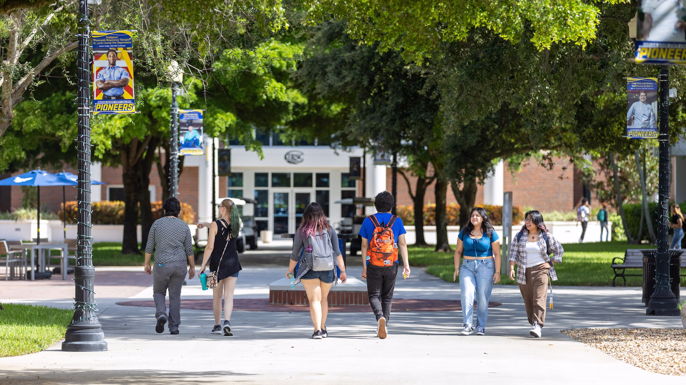 Students walk along a tree-lined pathway on a college campus with banners hanging from light poles on both sides of the walkway