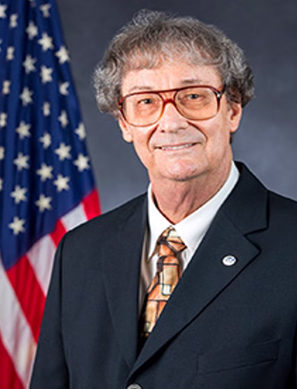 Professional headshot of Dr. Harvey Arnold in dark suit jacket and patterned tie with glasses, smiling in front of American flag