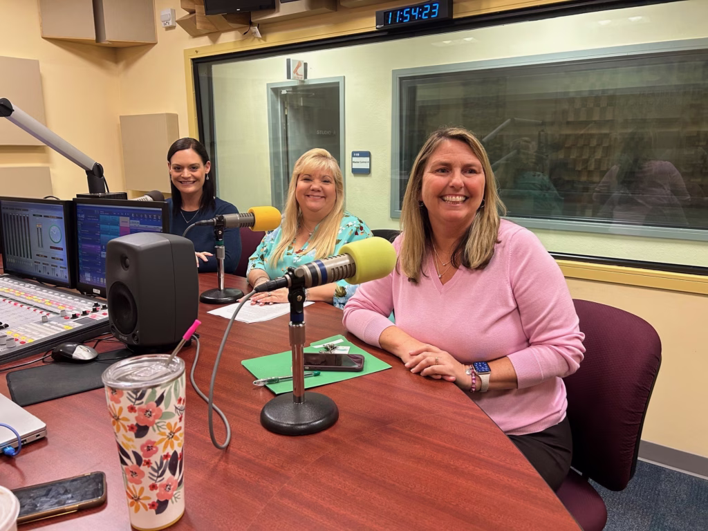 Three women seated at radio broadcast table with microphones, audio mixing board, and studio windows in background