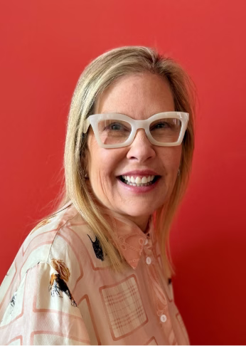 Woman with blonde hair and white-framed glasses smiling at camera against red background