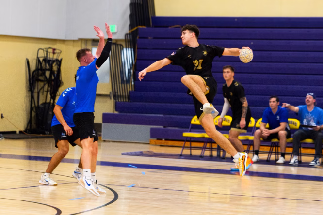 Handball player in black jersey number 21 leaps through the air holding a ball while a defender in blue raises his arms to block during an indoor gymnasium match