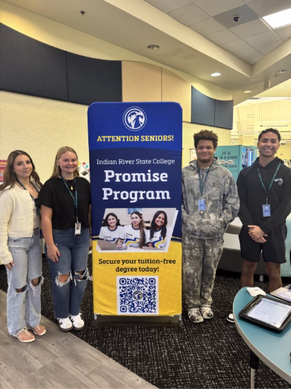 Four students standing beside a blue and yellow promotional banner for Indian River State College Promise Program. The banner reads "Attention Seniors!" at the top and "Secure your tuition-free degree today!" at the bottom, with a QR code. The students are wearing ID badges and casual clothing, positioned in what appears to be a school library or media center with yellow cabinets visible in the background.
