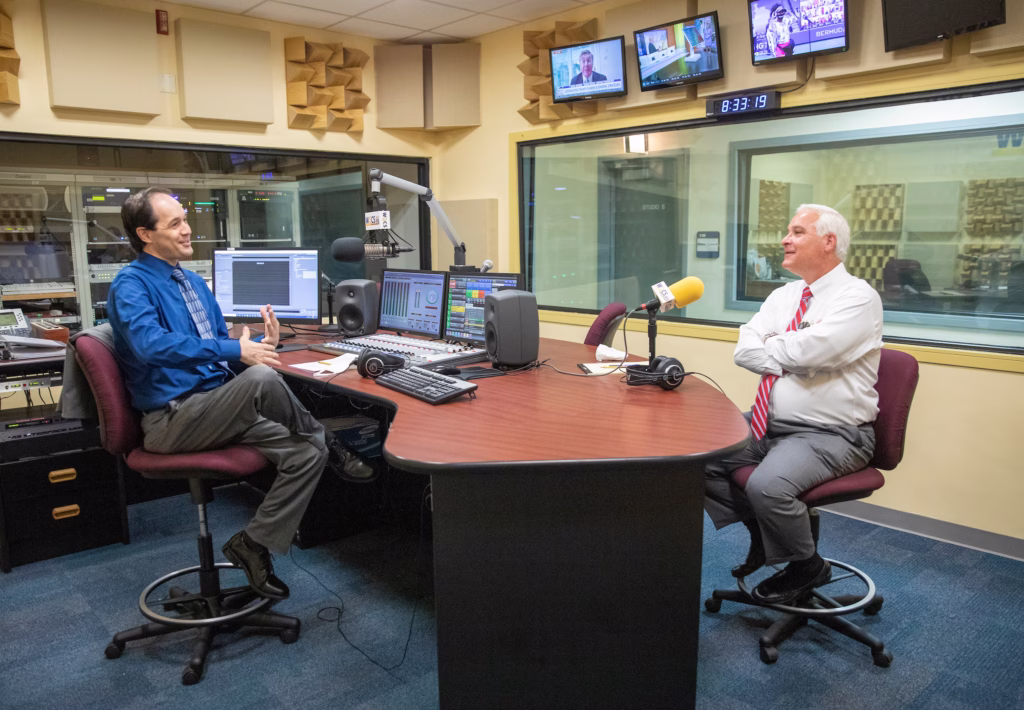 Two men conducting a radio interview in a professional broadcast studio, seated at a table with microphones, broadcasting equipment, and acoustic panels visible on the walls, with monitors displaying news broadcasts in the background.