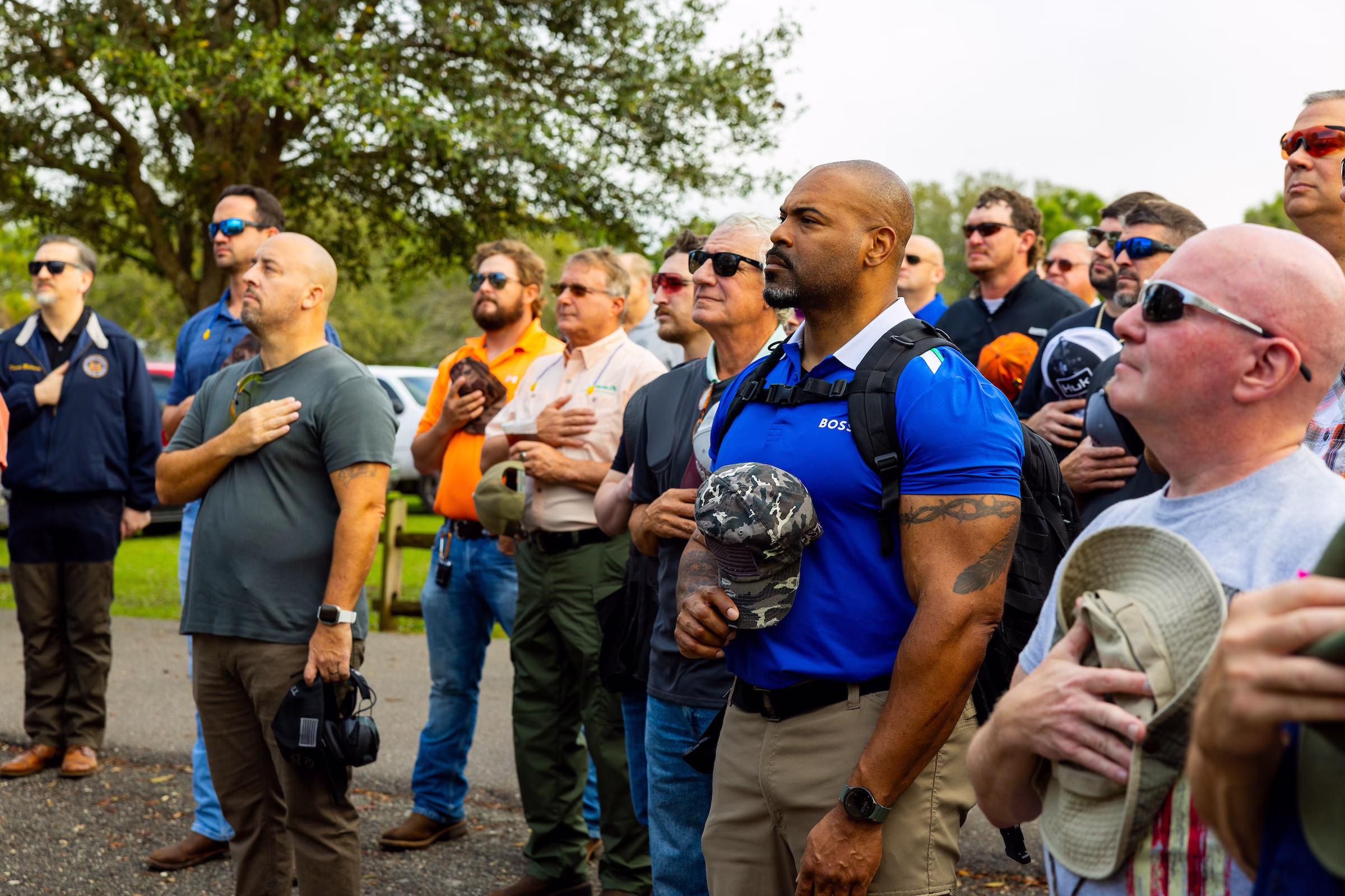 A group of people standing with hands over their hearts during the national anthem at a Veterans Day Clay Shootout event, surrounded by trees in an outdoor setting.