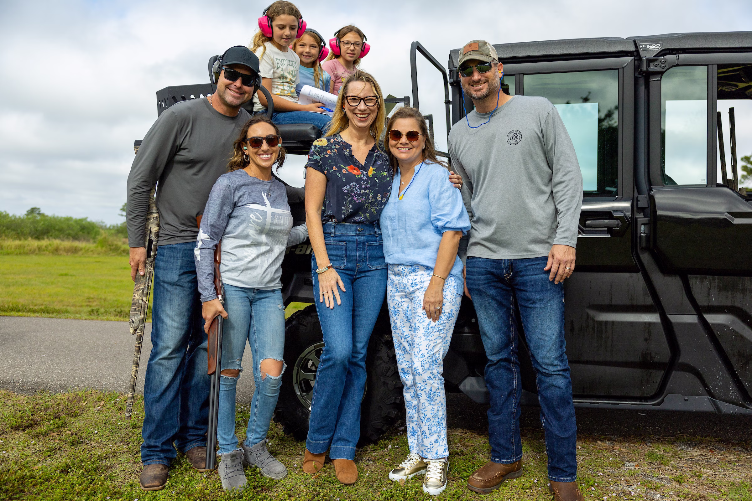 A multigenerational family of seven poses in front of a black off-road vehicle at an outdoor clay shooting event. Four adults stand in front while three young girls wearing bright pink ear protection sit on top of the vehicle's roll bar. One adult holds a shotgun. The group is smiling at the camera with open fields visible in the background.