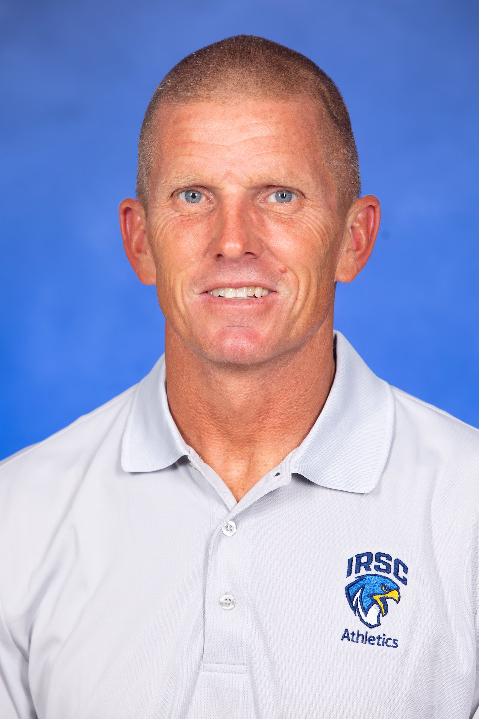 Headshot of Sion Brinn, head swimming and diving coach at Indian River State College, smiling against a blue background while wearing a white IRSC Athletics polo shirt.