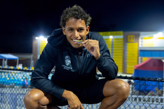 A young male swimmer in a black IRSC Swimming & Diving jacket crouches outdoors at night, smiling and biting a gold medal.