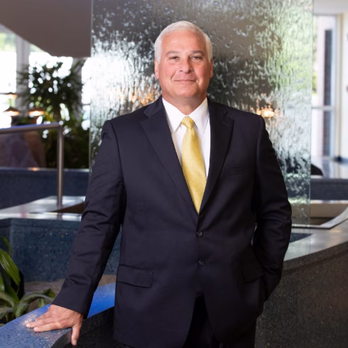 Professional headshot of a man in a dark suit and gold tie, standing in a modern lobby with a water feature and greenery in the background.
