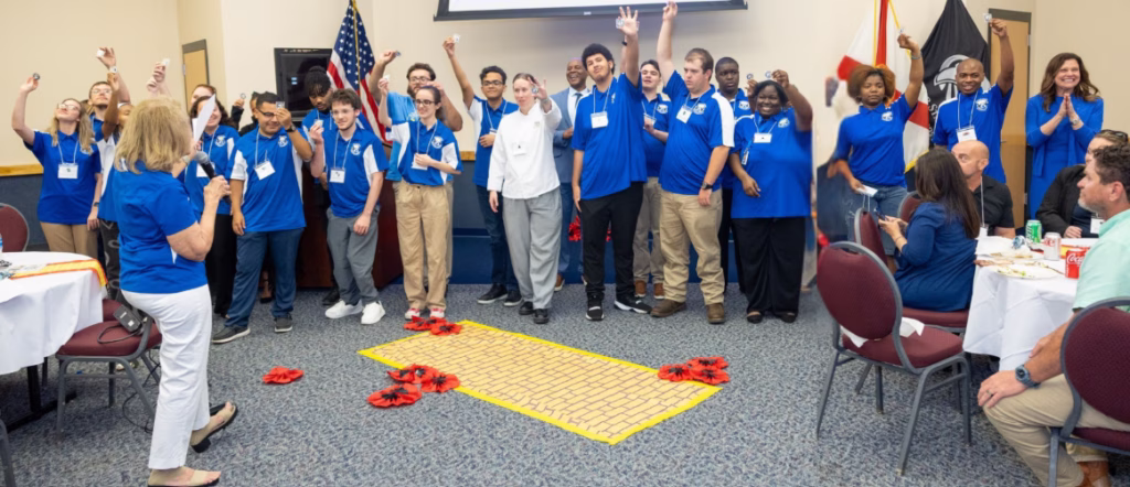 A large group of students in blue polo shirts celebrate with raised hands and cheers in a conference room, standing behind a yellow brick road decoration with red poppy flowers on the floor, as a woman with a microphone addresses them.