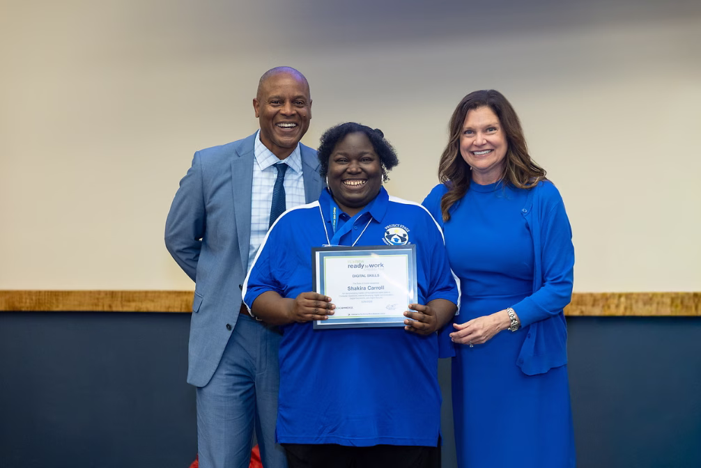 A woman in a blue polo shirt smiles while holding a certificate at an awards ceremony, flanked by two presenters — a man in a grey suit and a woman in a blue dress.