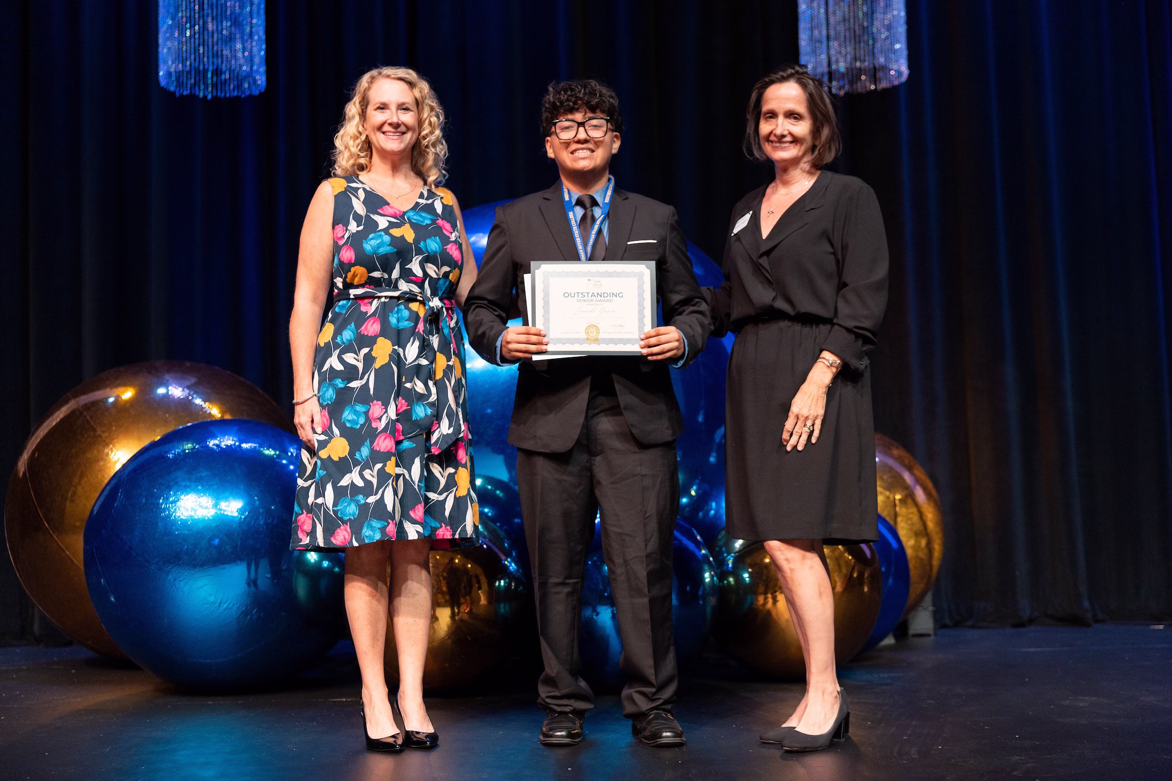A young male student wearing a black suit, tie, glasses, and a medal stands center stage between two women, proudly holding an Outstanding Senior Award certificate. The woman on the left wears a floral dress; the woman on the right wears a black dress. Large blue and gold metallic balloons and blue draping decorate the stage behind them.