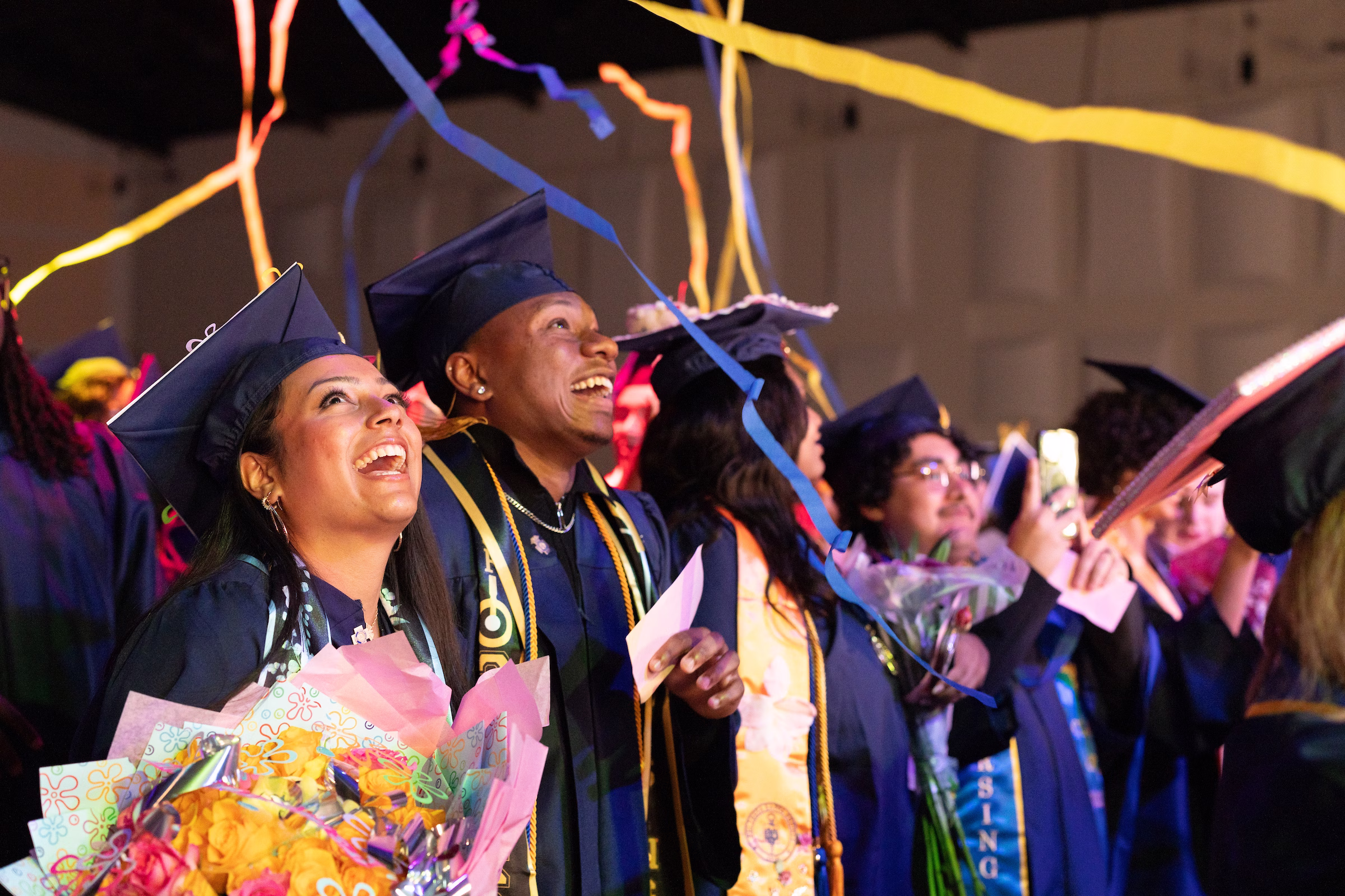 A group of nursing graduates in navy blue caps and gowns laugh and look upward as colorful streamers fall around them during a commencement ceremony. The graduate in the foreground holds a bouquet of yellow and pink flowers, while others hold diplomas and capture the moment on their phones.