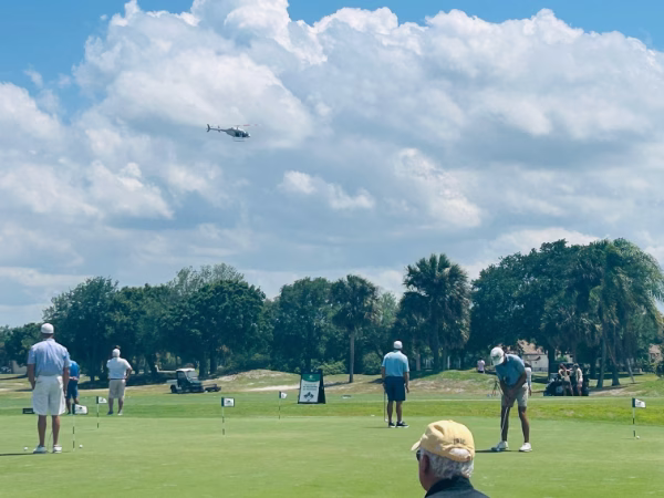 Golfers stand on a sunny Florida golf course fairway surrounded by palm trees beneath a partly cloudy blue sky. A helicopter flies overhead in the background. A tournament sign is visible near the tree line.