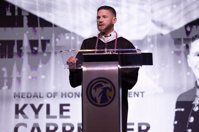 Medal of Honor recipient Kyle Carpenter speaks at a podium during a commencement ceremony, wearing academic regalia. A projection screen behind him displays his name and title.