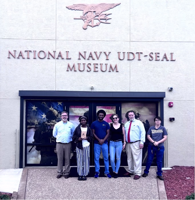 Six people stand together in front of the National Navy UDT-SEAL Museum entrance in Fort Pierce, Florida. The museum's name and military emblem are visible on the building behind them.