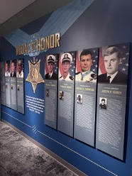 A wide museum display wall titled "Medal of Honor" features large portrait panels and biographical descriptions of Navy SEAL recipients, set against a blue background with gold lettering.