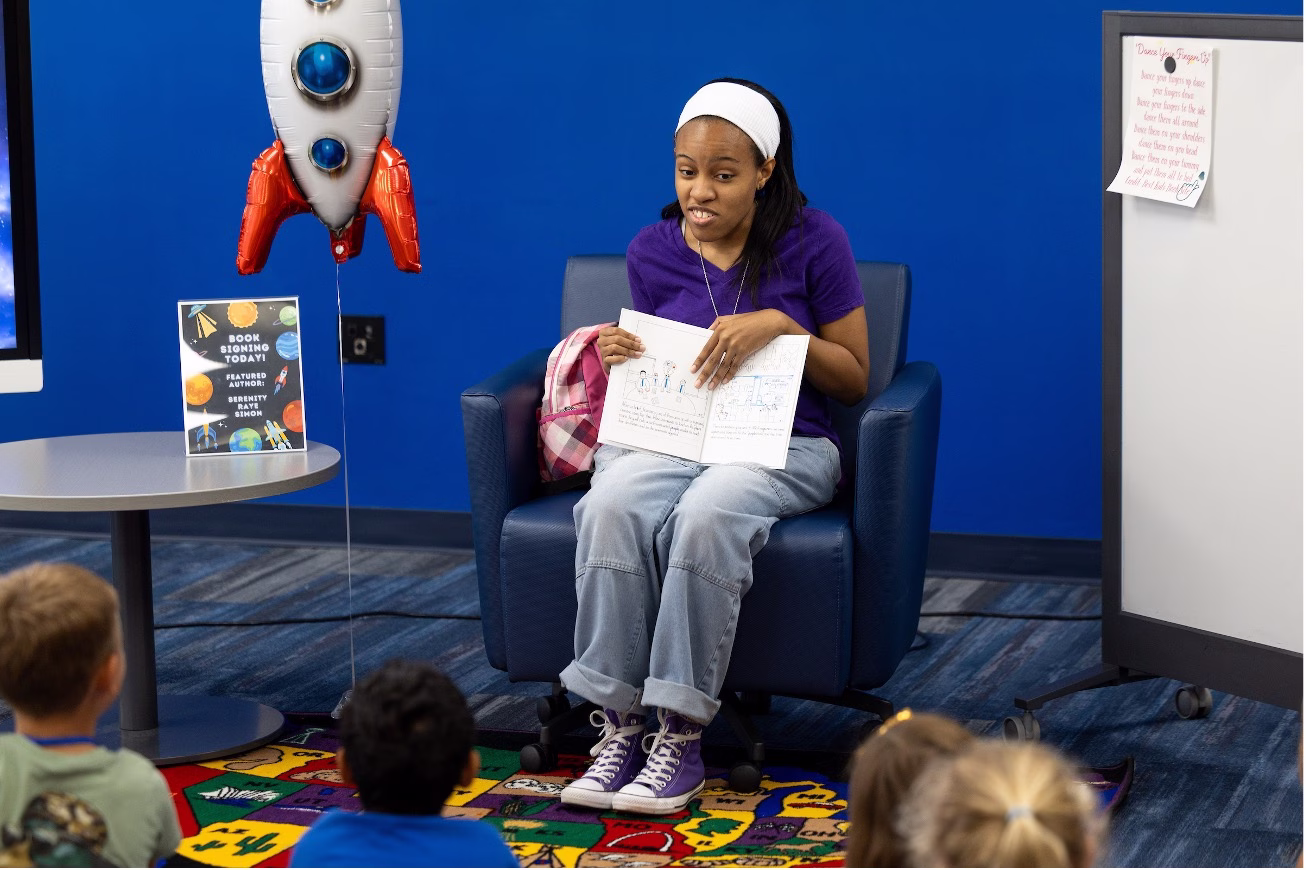 A young woman in a purple shirt and white headband sits in a blue armchair reading an open children's book to a group of young children seated on a colorful rug. A rocket-shaped balloon and a "Book Signing Today — Featured Author: Serenity Raye Simon" sign are visible nearby.
