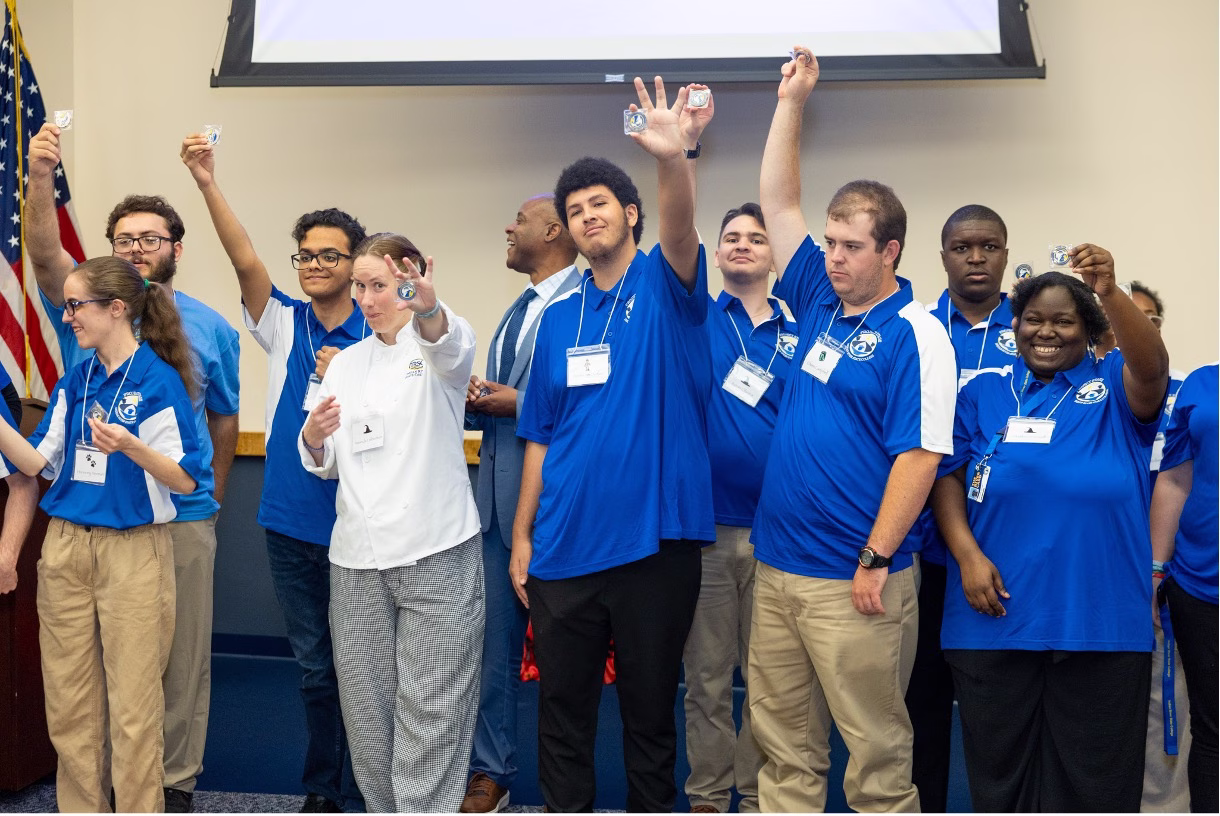 A group of Project STAGE students wearing blue and white polo shirts hold up small challenge coins at the program's 2nd Annual Banquet. A faculty member in a white chef's coat stands among them, and an American flag is visible in the background.